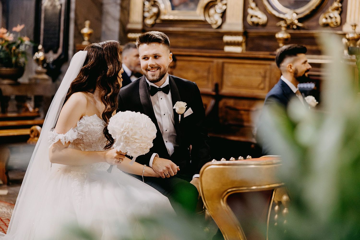 Smiling groom in tuxedo looking at bride holding white bouquet during church wedding ceremony.