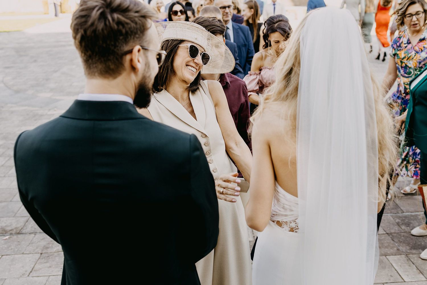 Bride and groom share a moment with a smiling guest wearing a sun hat and sunglasses at an outdoor wedding.