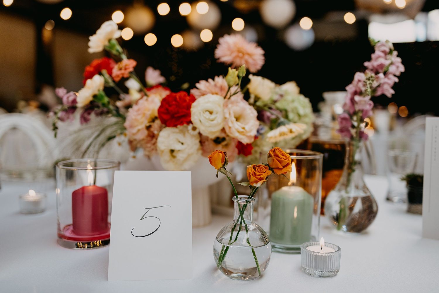 Colorful wedding table centerpiece with pink roses, candles, and a number five card on white tablecloth.