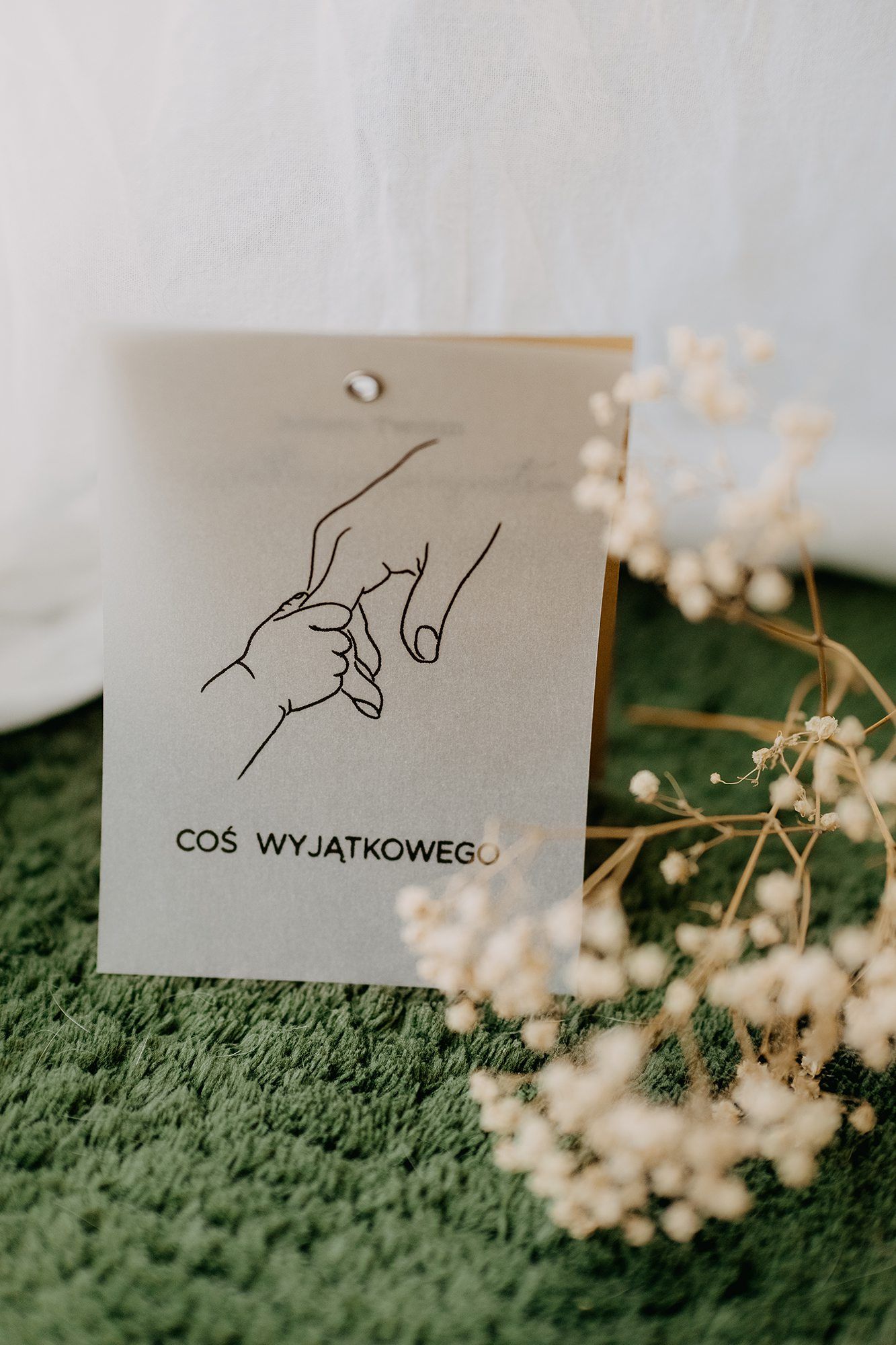A wedding gift tag reading 'Cos Wyjatkowego' with a hand-drawn couple illustration, surrounded by delicate white dried flowers.