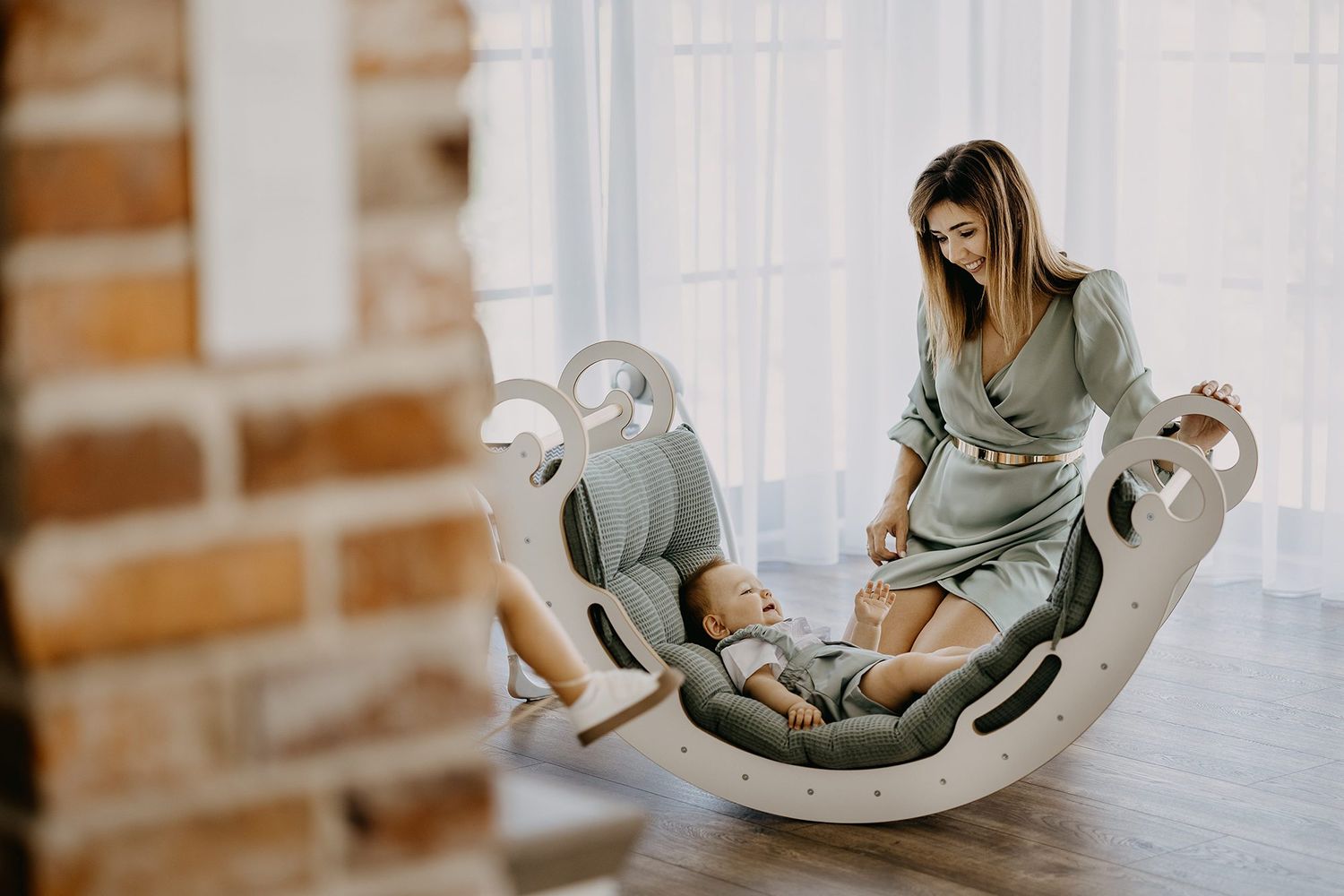 Young mother in sage dress sitting with smiling baby on wooden rocking toy in bright room.