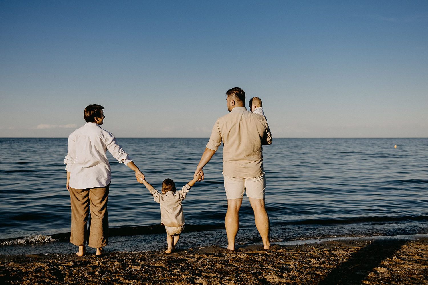 Two dads and children stand hand-in-hand facing the calm ocean on a sunny beach day.