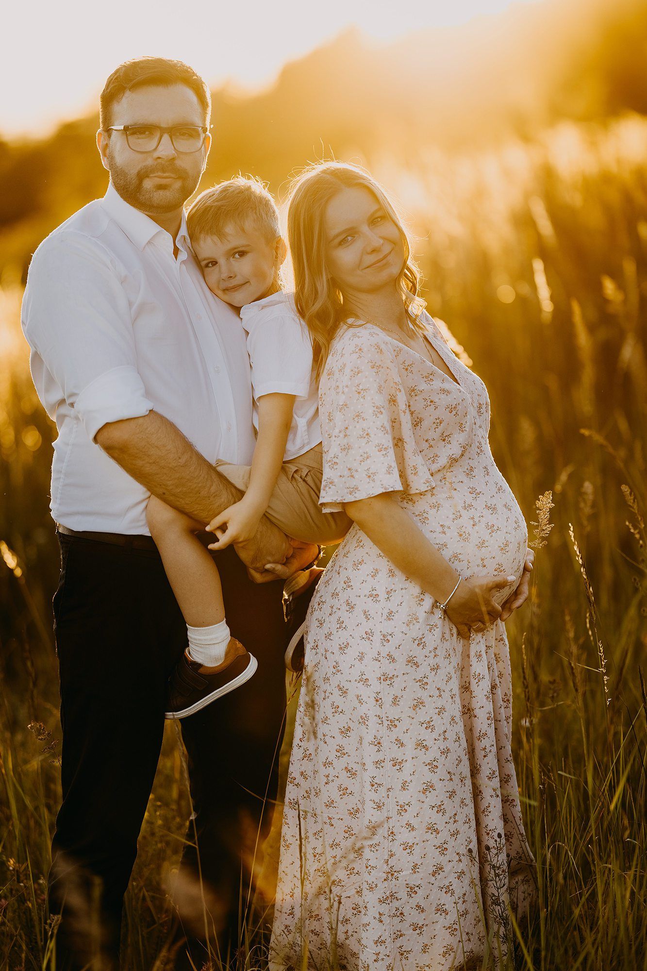 Happy family with young son posing in golden sunset field during outdoor maternity photo session.