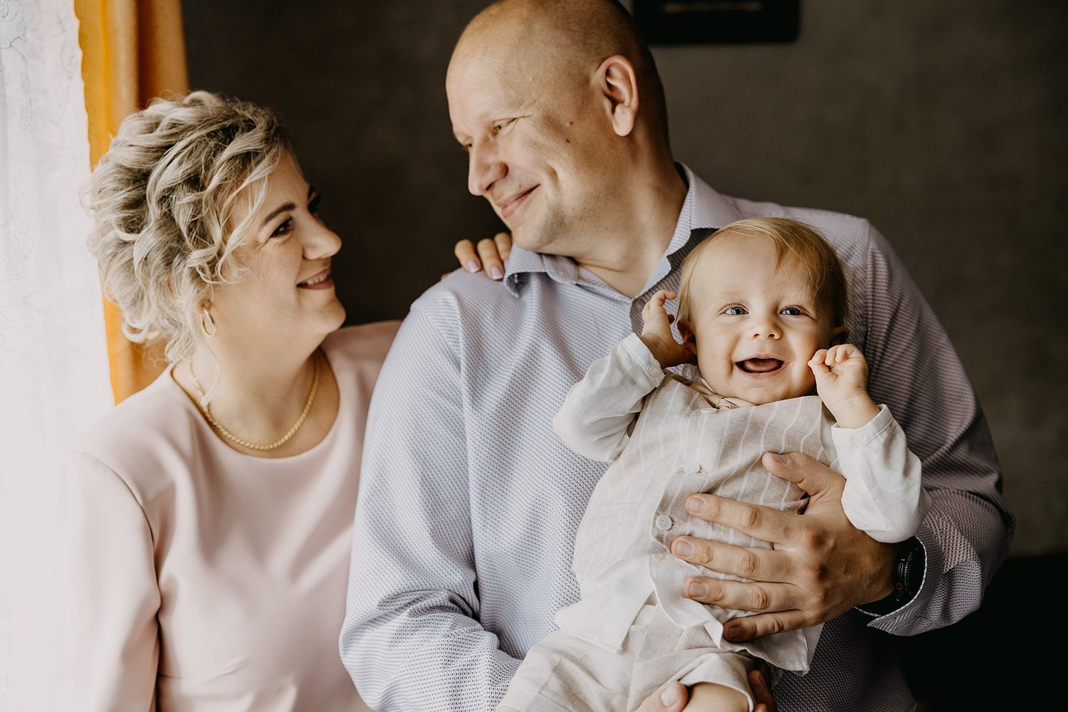 Happy family portrait with smiling parents holding a laughing baby near a bright window.