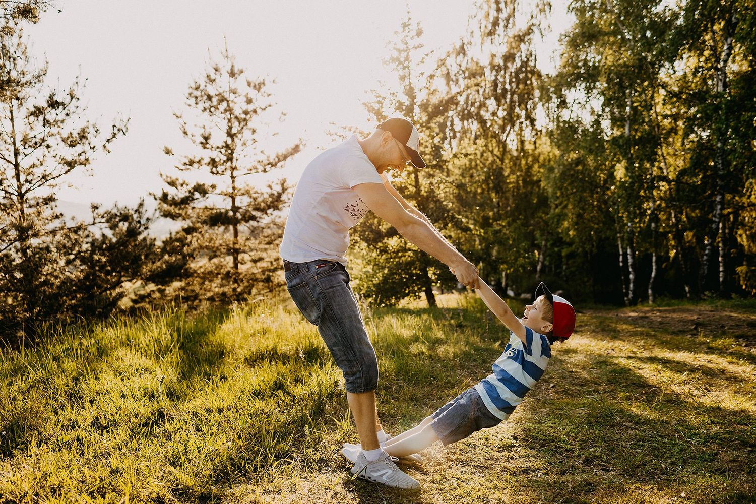 A father swings his young child by the arms in a sunny golden forest clearing.