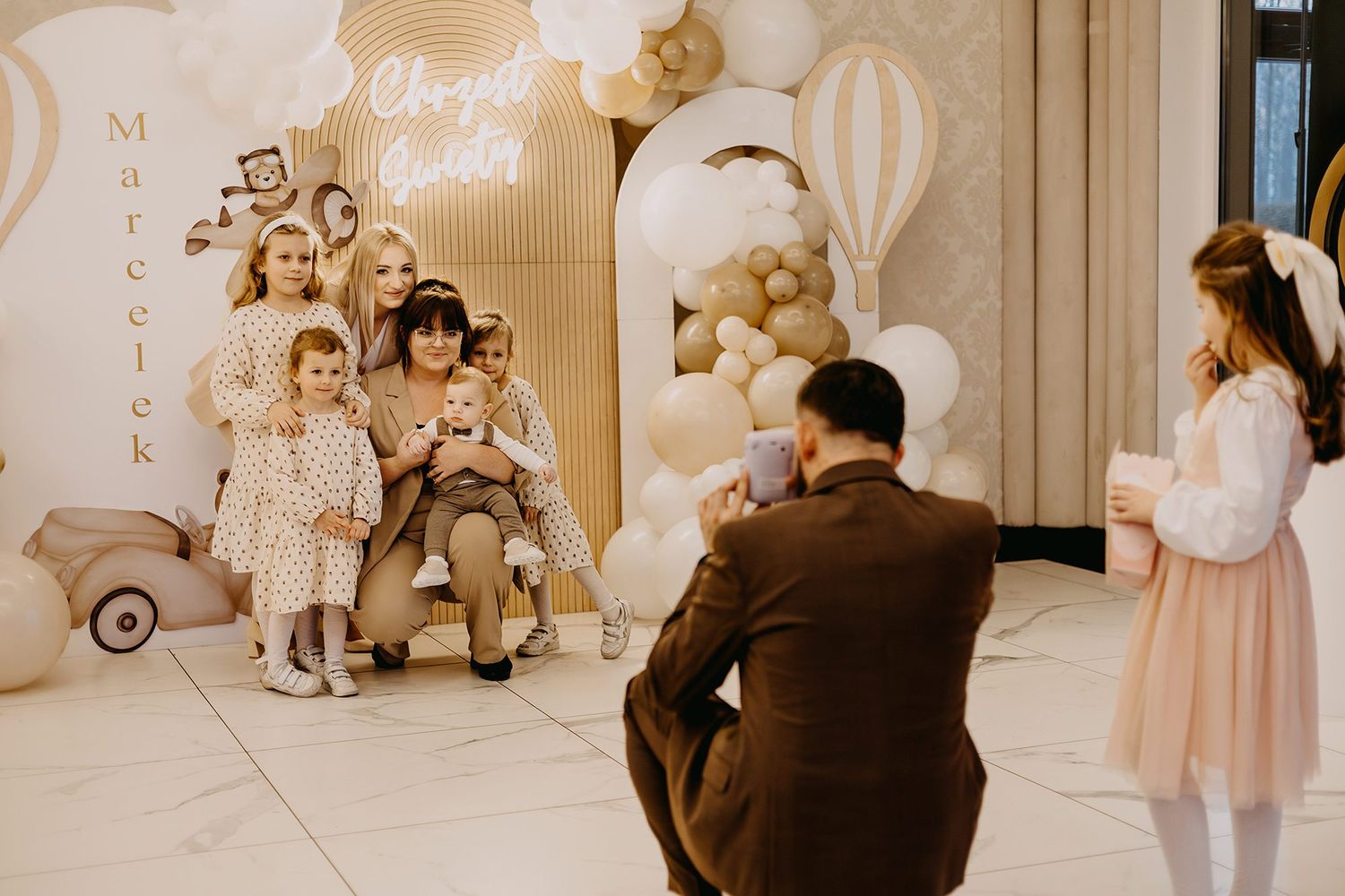 Family posing for photos at a christening party in front of a decorated balloon backdrop display.