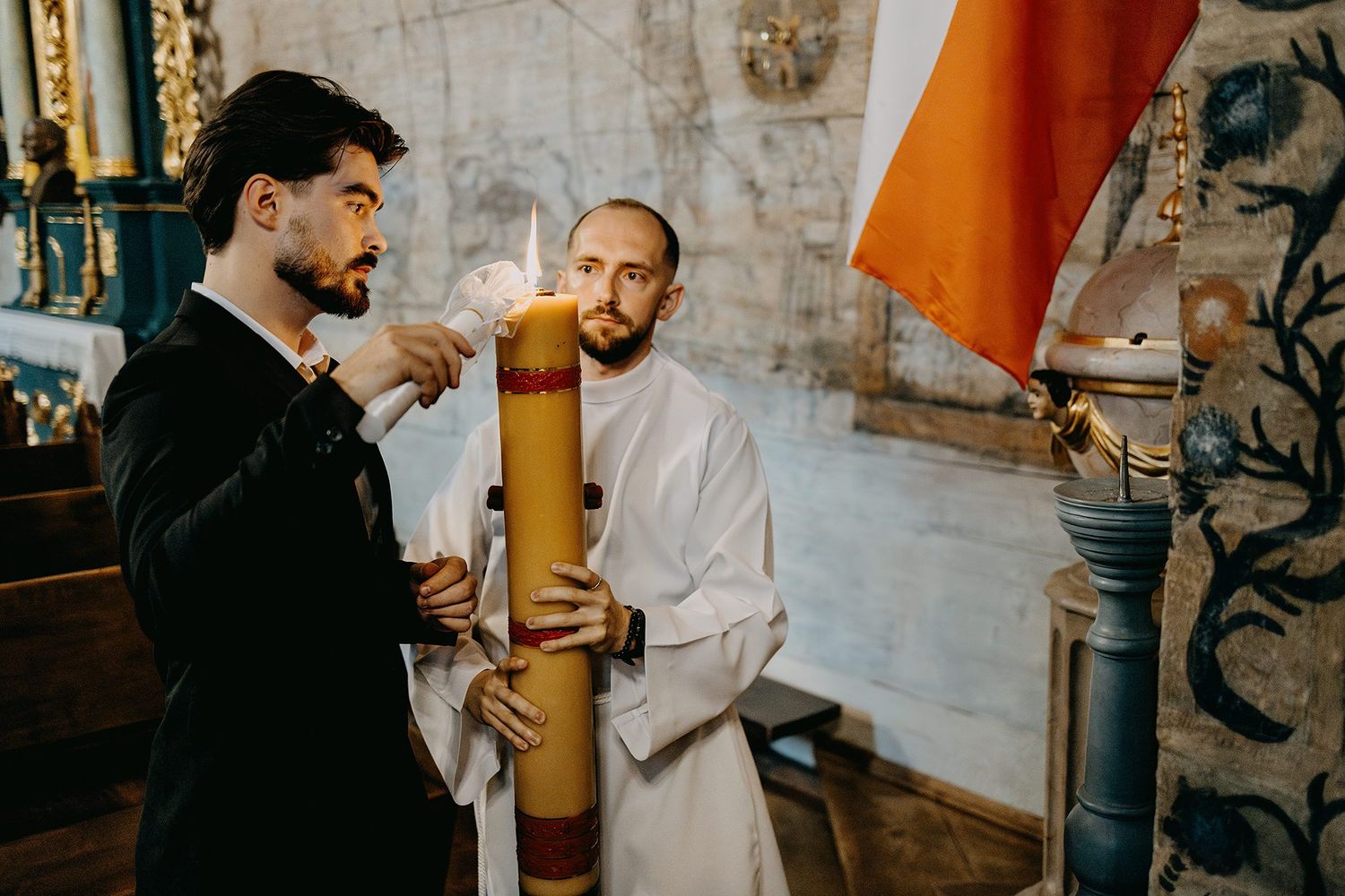 A godfather lights a large baptismal candle assisted by a priest during a christening.