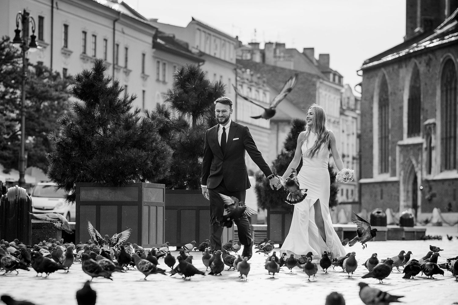 Bride and groom walk through a city square surrounded by pigeons in a black and white wedding photo.