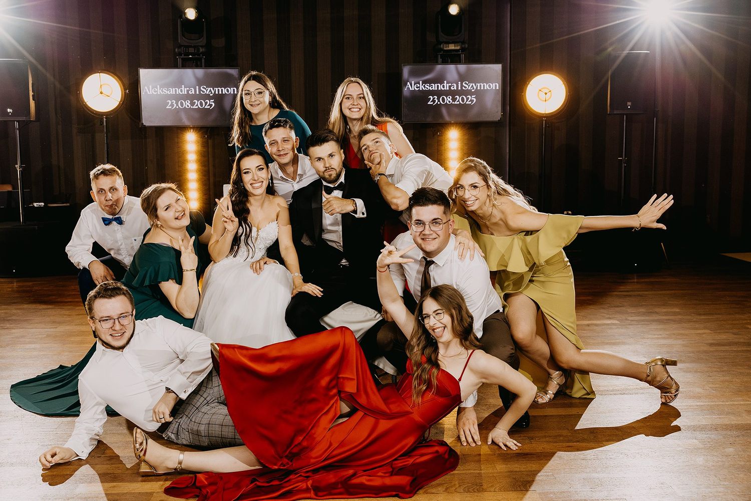 Joyful wedding party group photo with bride, groom, and friends posing playfully on a dance floor.