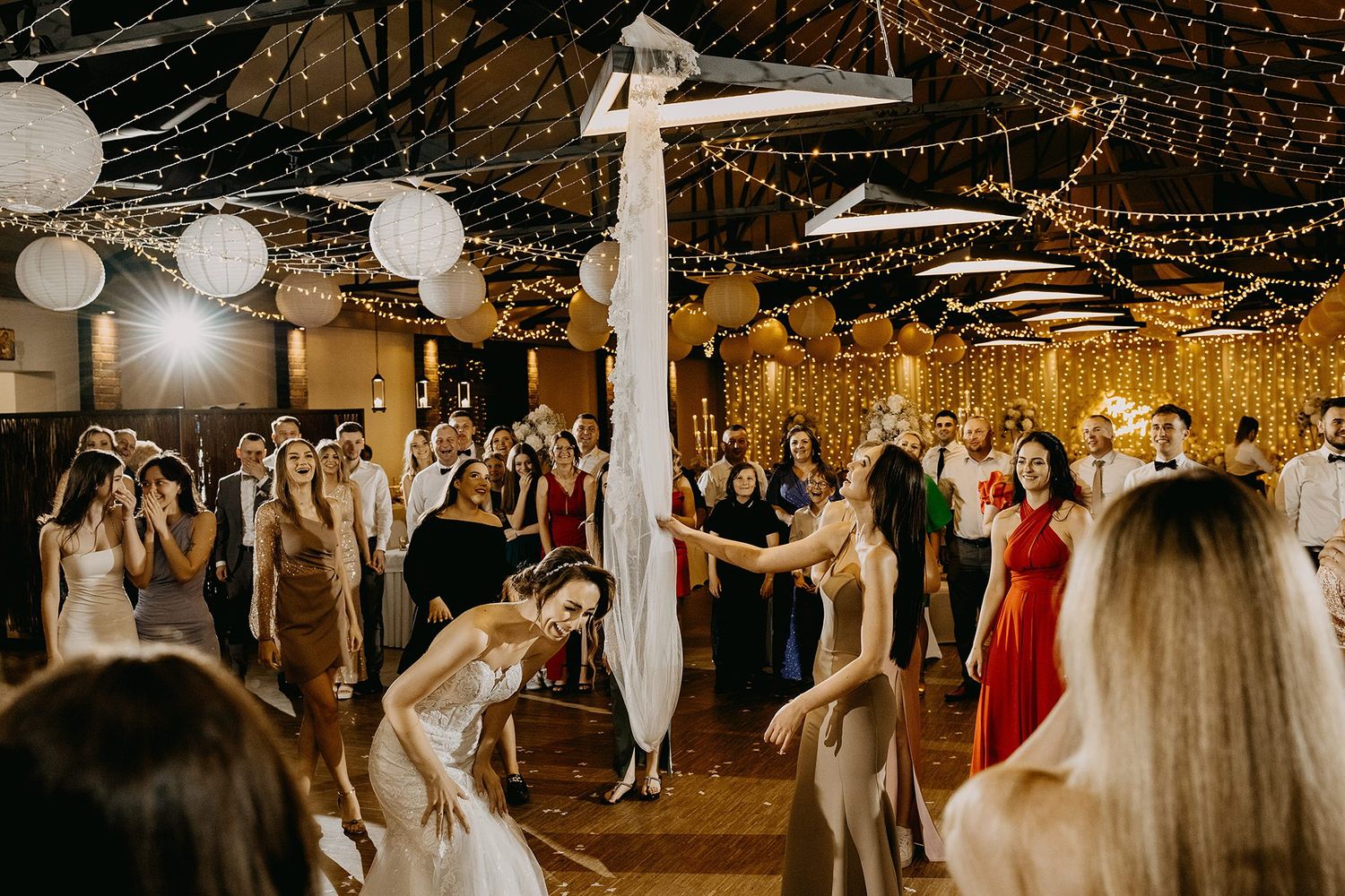 Bride tossing bouquet to excited guests on decorated wedding reception dance floor with string lights.