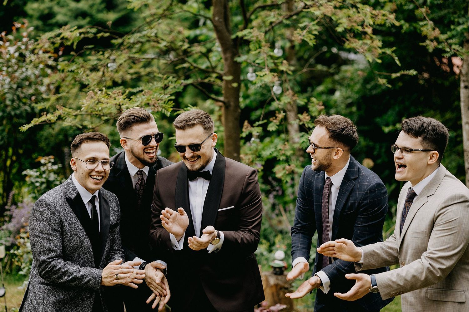 Groom and groomsmen laughing together outdoors in suits at a wedding celebration.