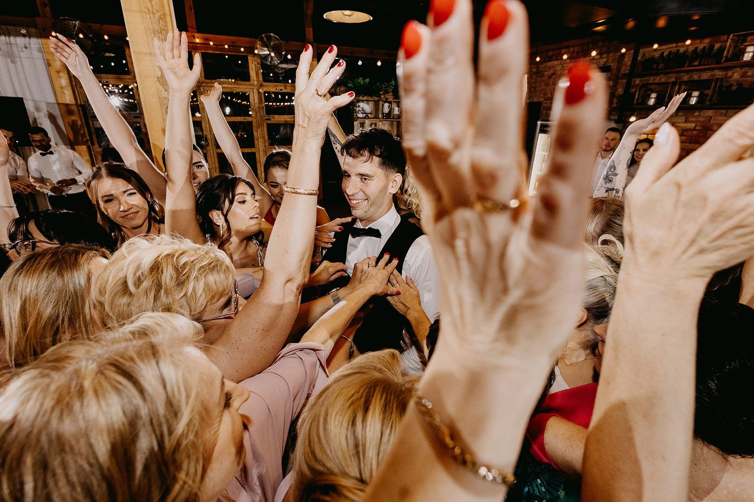 Energetic wedding DJ surrounded by enthusiastic guests with raised hands on the dance floor.