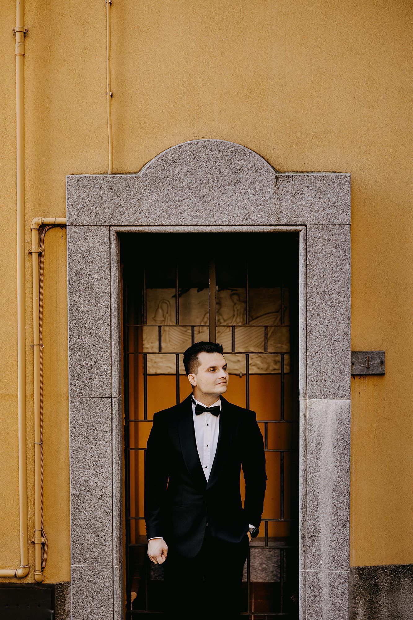 Dapper groom in black tuxedo stands confidently in a decorative doorway against a yellow wall.
