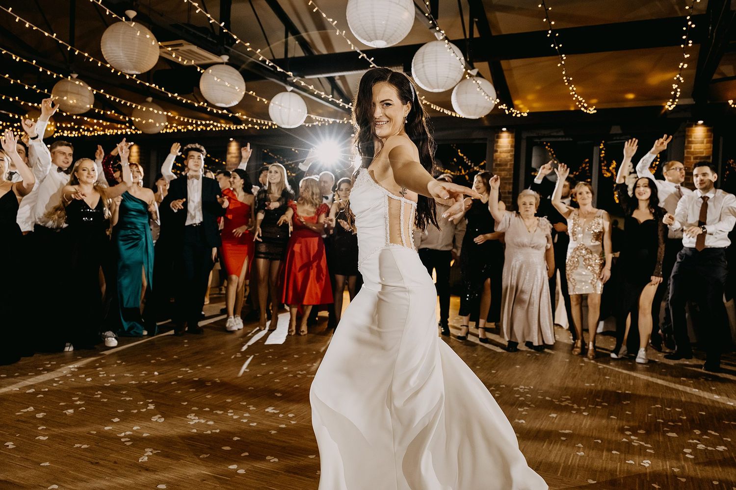 Radiant bride in white gown twirls on dance floor while wedding guests celebrate with sparklers behind her.