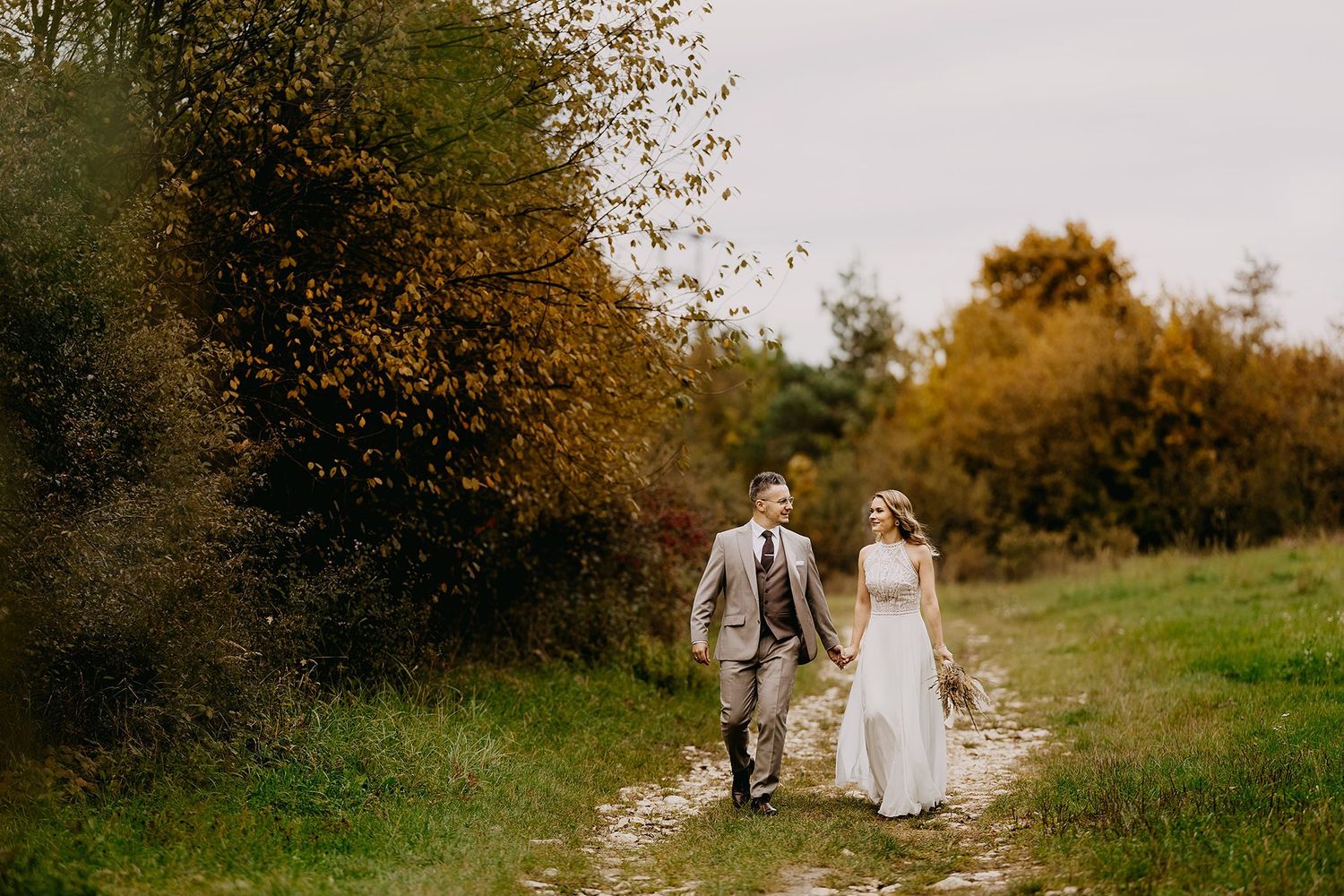 Newlyweds walking hand in hand along a countryside path surrounded by autumn foliage.
