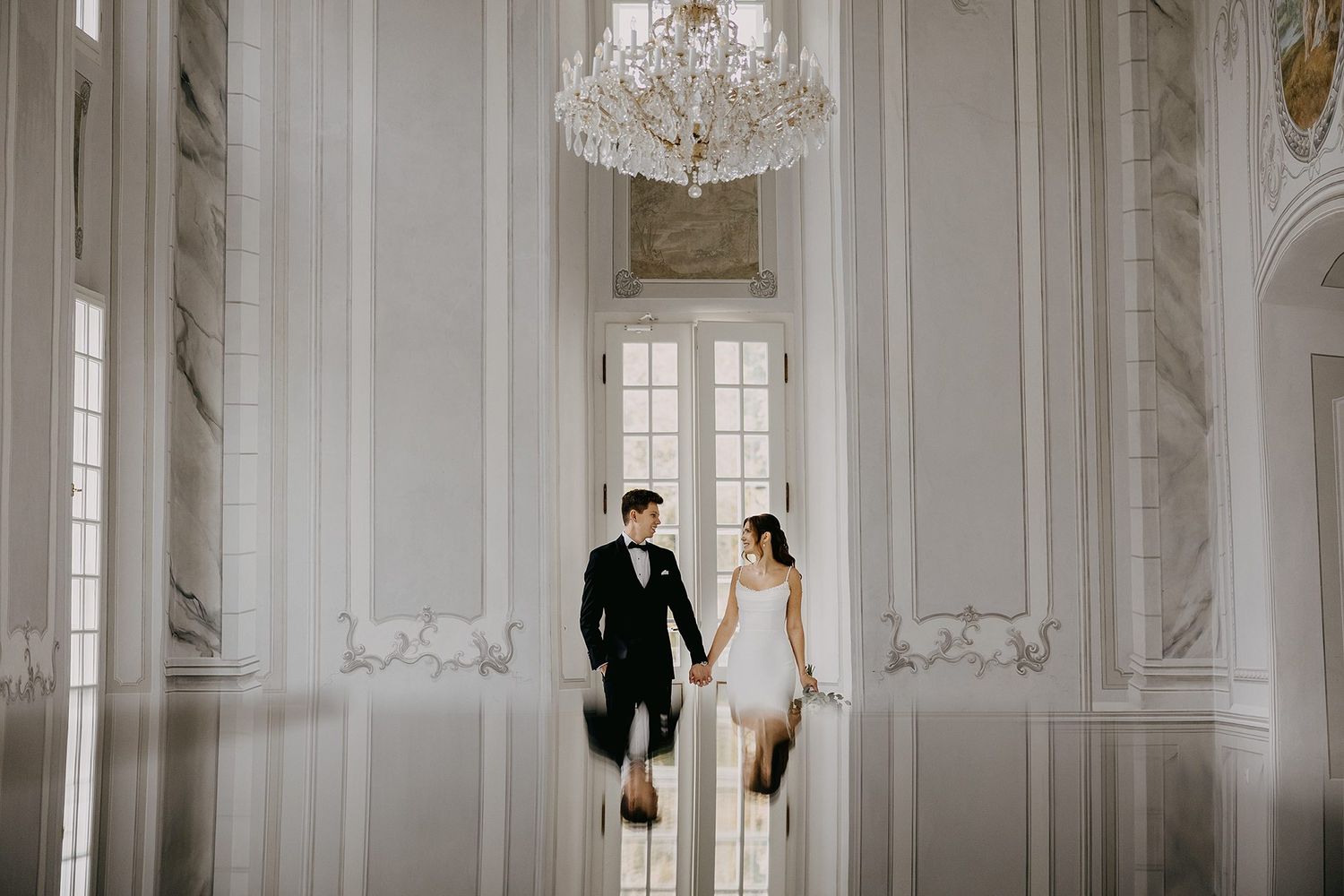 Bride and groom walking hand in hand in an elegant white ballroom with chandelier.