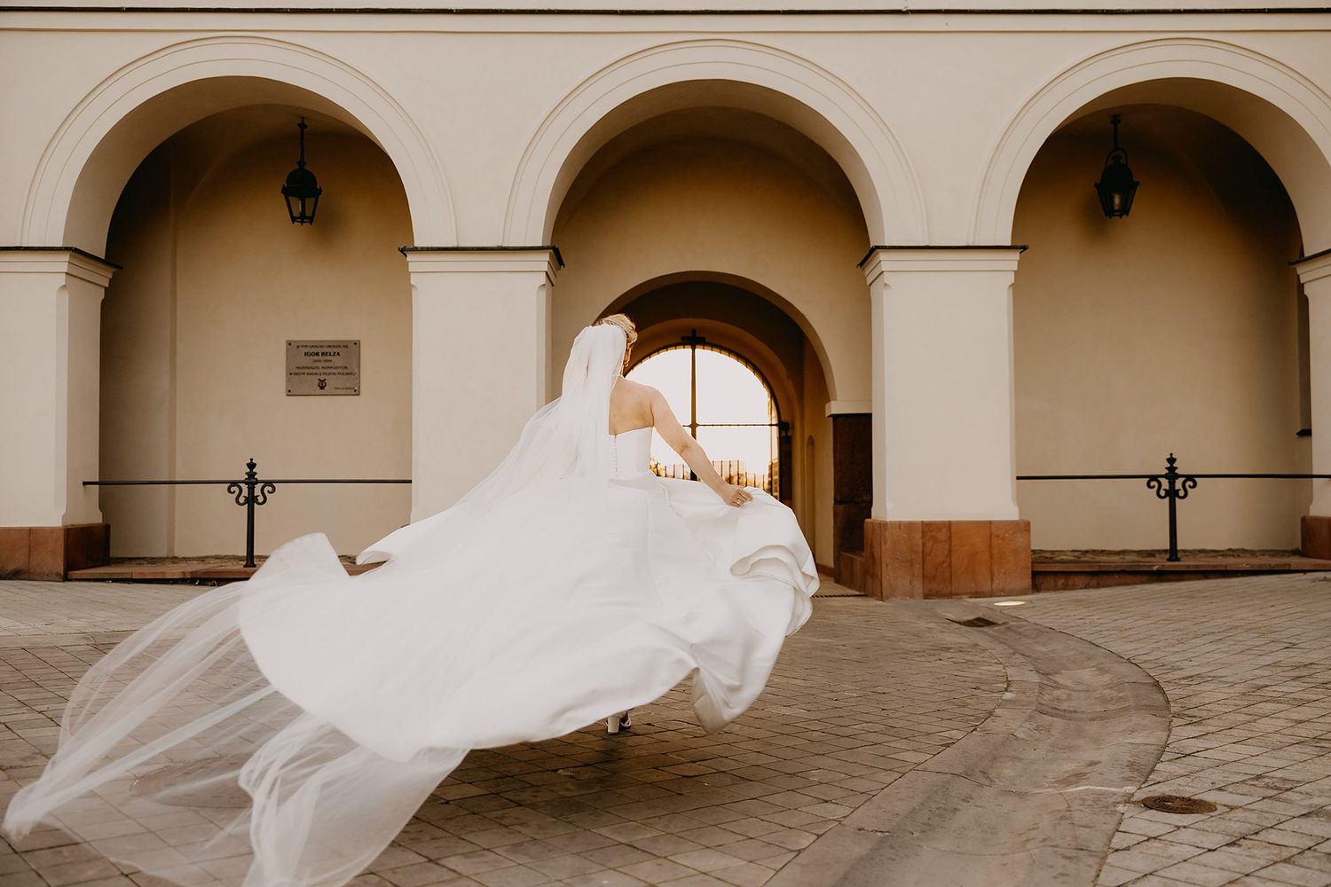 Bride twirling in flowing white wedding gown creating dramatic veil movement in front of arched building.