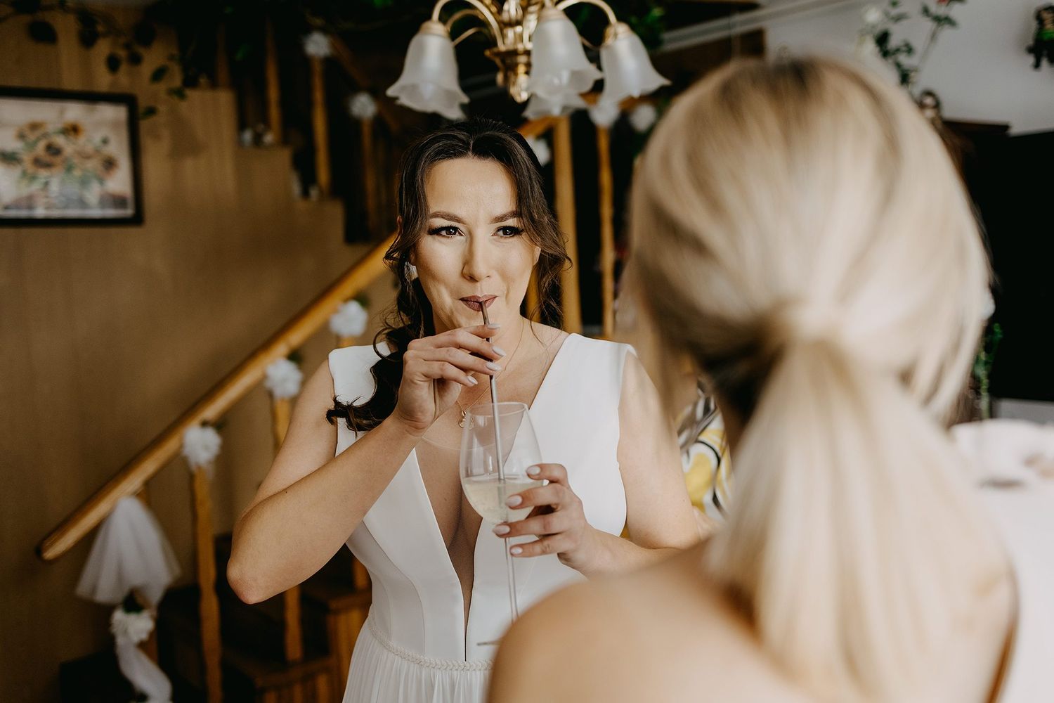 Bride in white dress sipping champagne with bridesmaid during wedding preparation indoors.