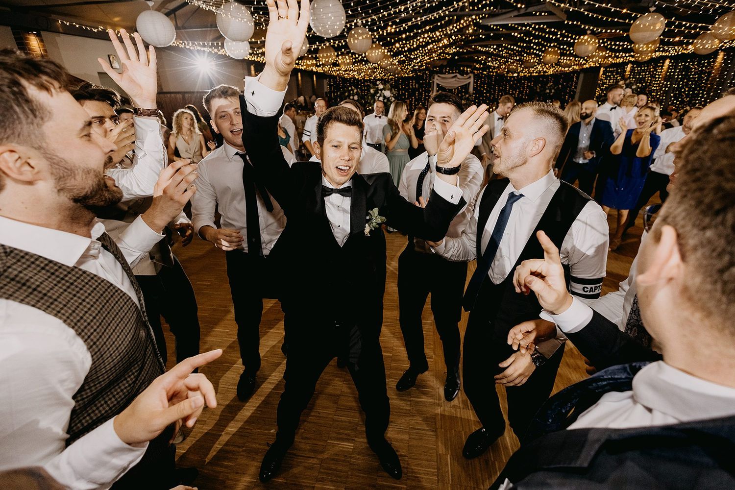 Groom celebrating with groomsmen on the dance floor at a lively wedding reception under string lights.