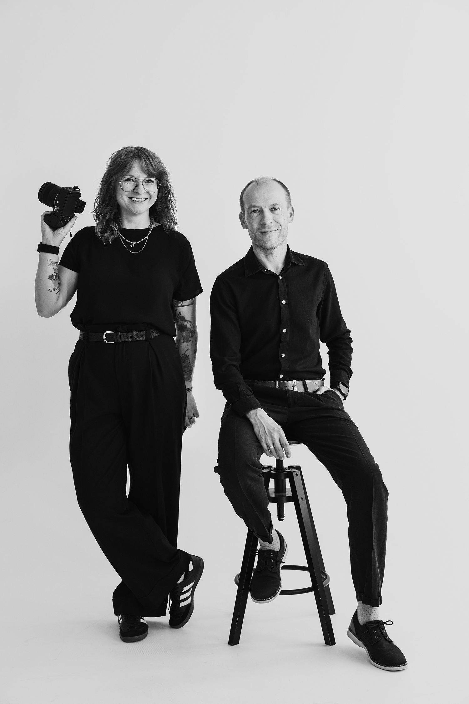 Black and white photo of two photographers in black outfits, woman holding camera standing beside man seated on stool.