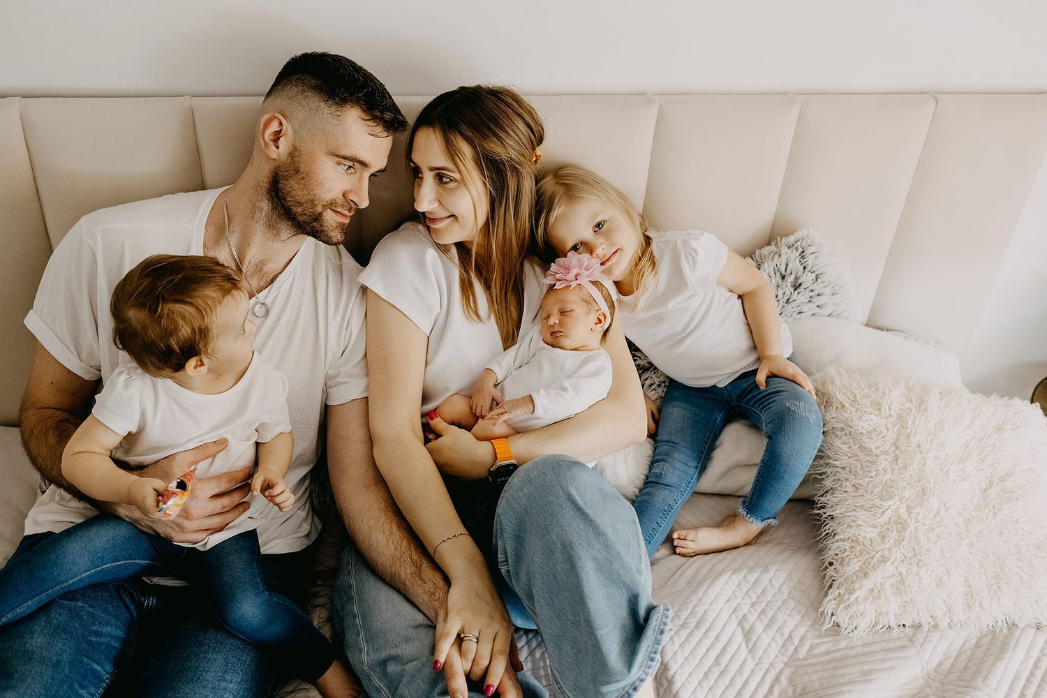 Happy family with three children sitting together on a bed, parents smiling at each other while holding their kids.