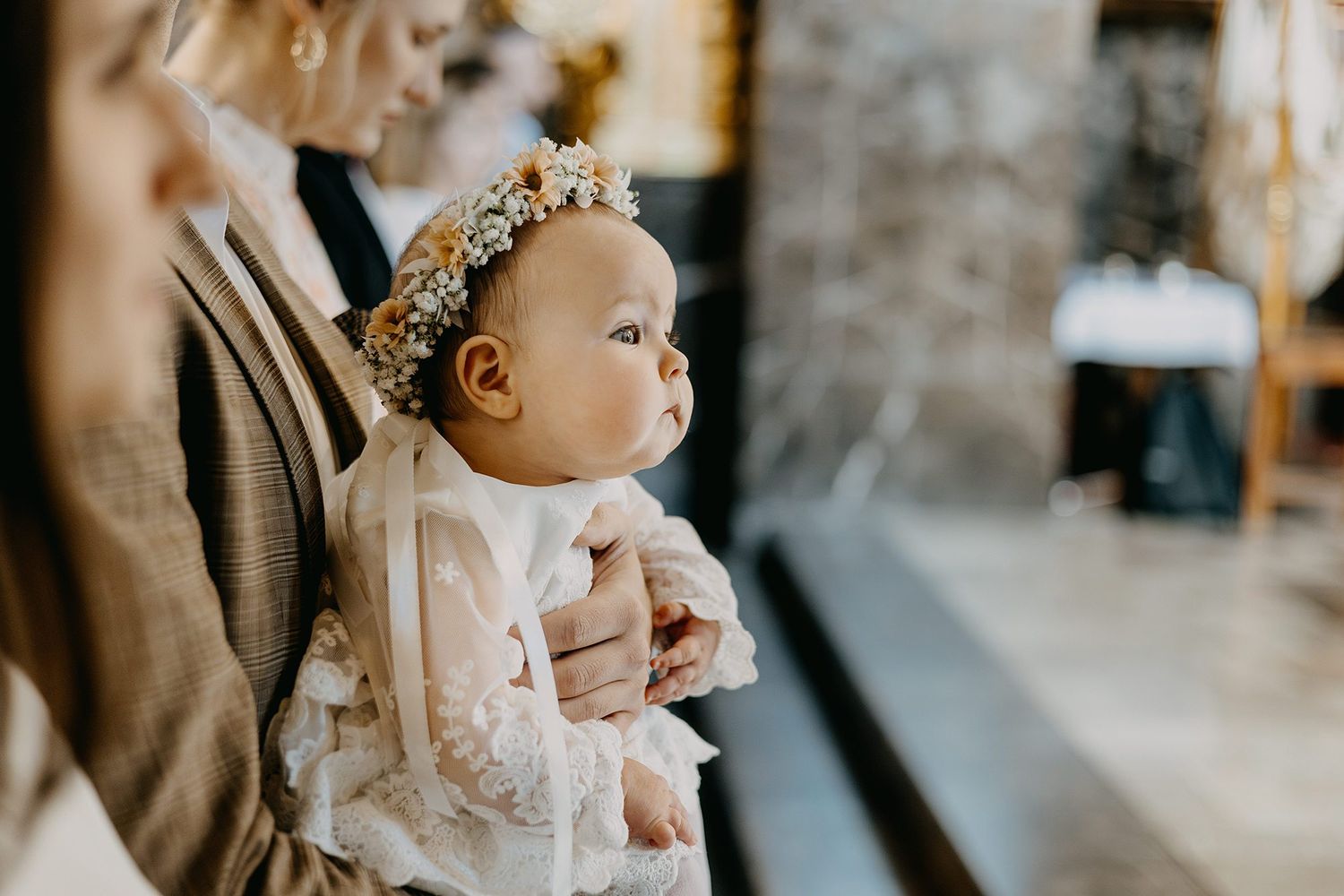 Baby girl wearing a floral crown in a white christening gown held during a church ceremony.