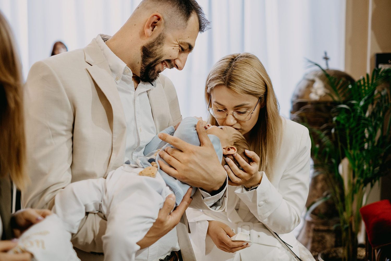A smiling couple in white outfits tenderly holds and kisses their newborn baby during a celebration.