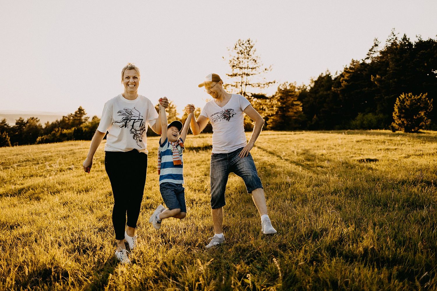 Happy family swinging young son by his hands in a sunlit green meadow.