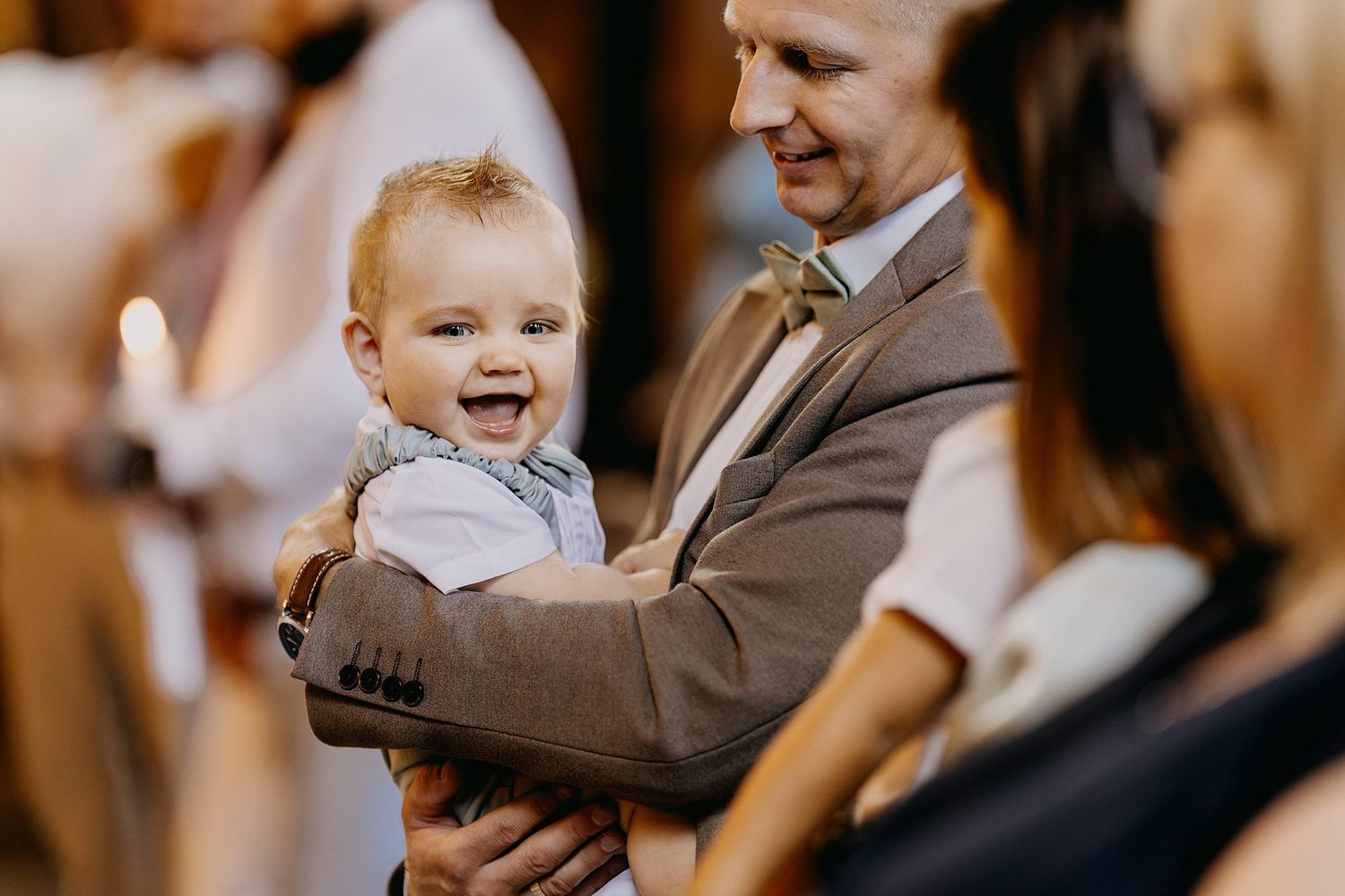 Smiling baby in white outfit held by man in gray suit at a church ceremony.