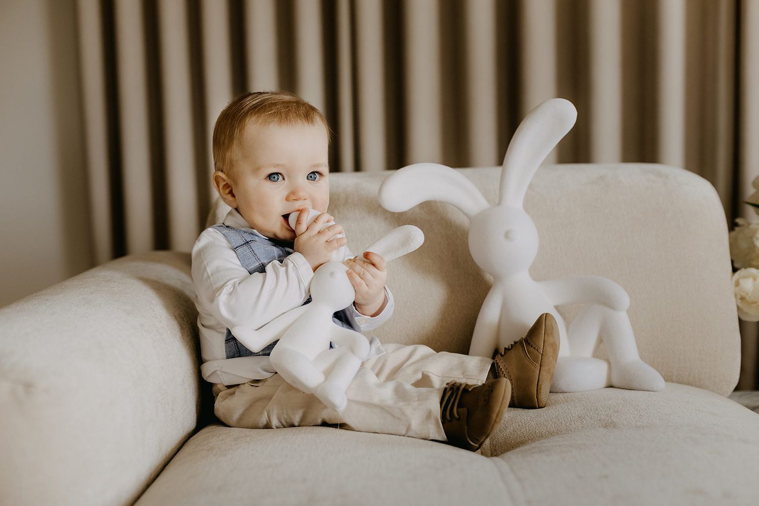An adorable baby in formal attire sits on a sofa beside a white bunny toy.