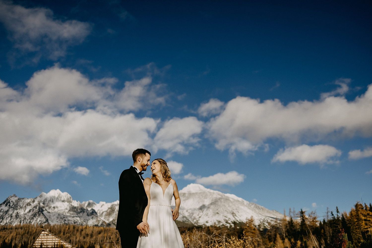 A bride and groom share a romantic moment with snow-capped mountains and blue sky in the background.