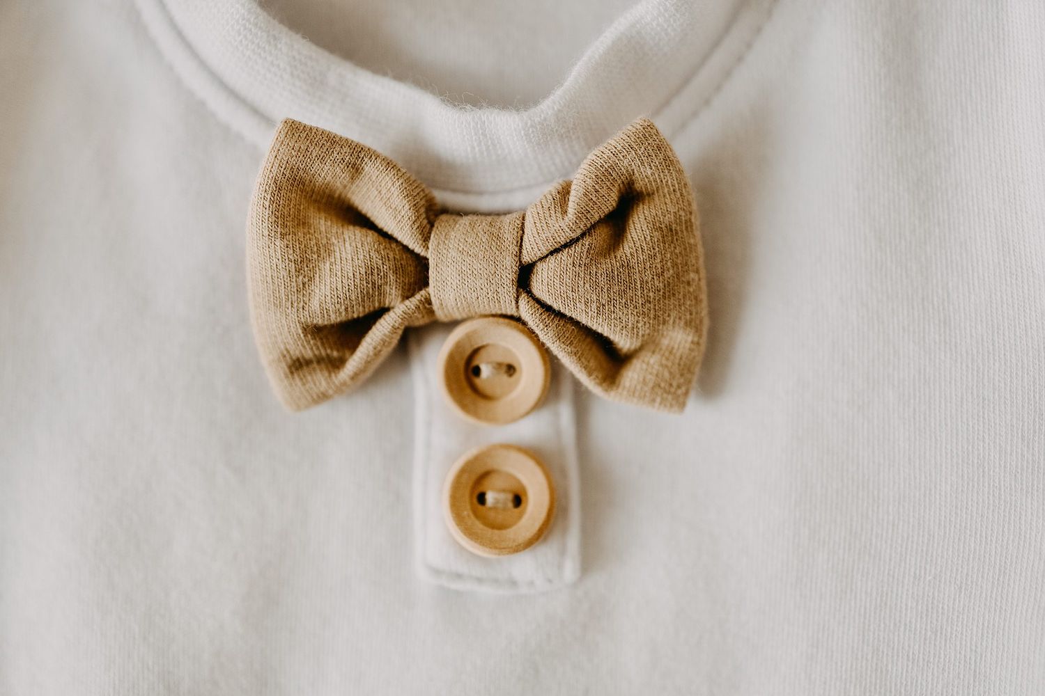 Close-up of a white baby onesie with a beige linen bow tie and wooden buttons detail.
