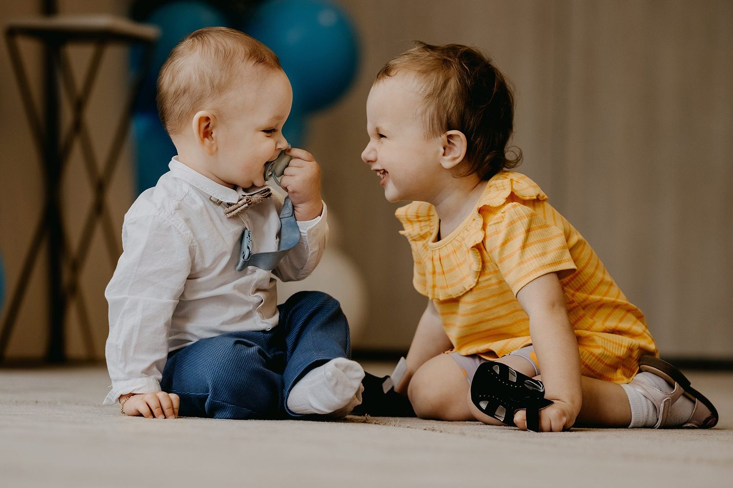 Two babies sit facing each other on the floor, smiling and interacting playfully during a birthday celebration.