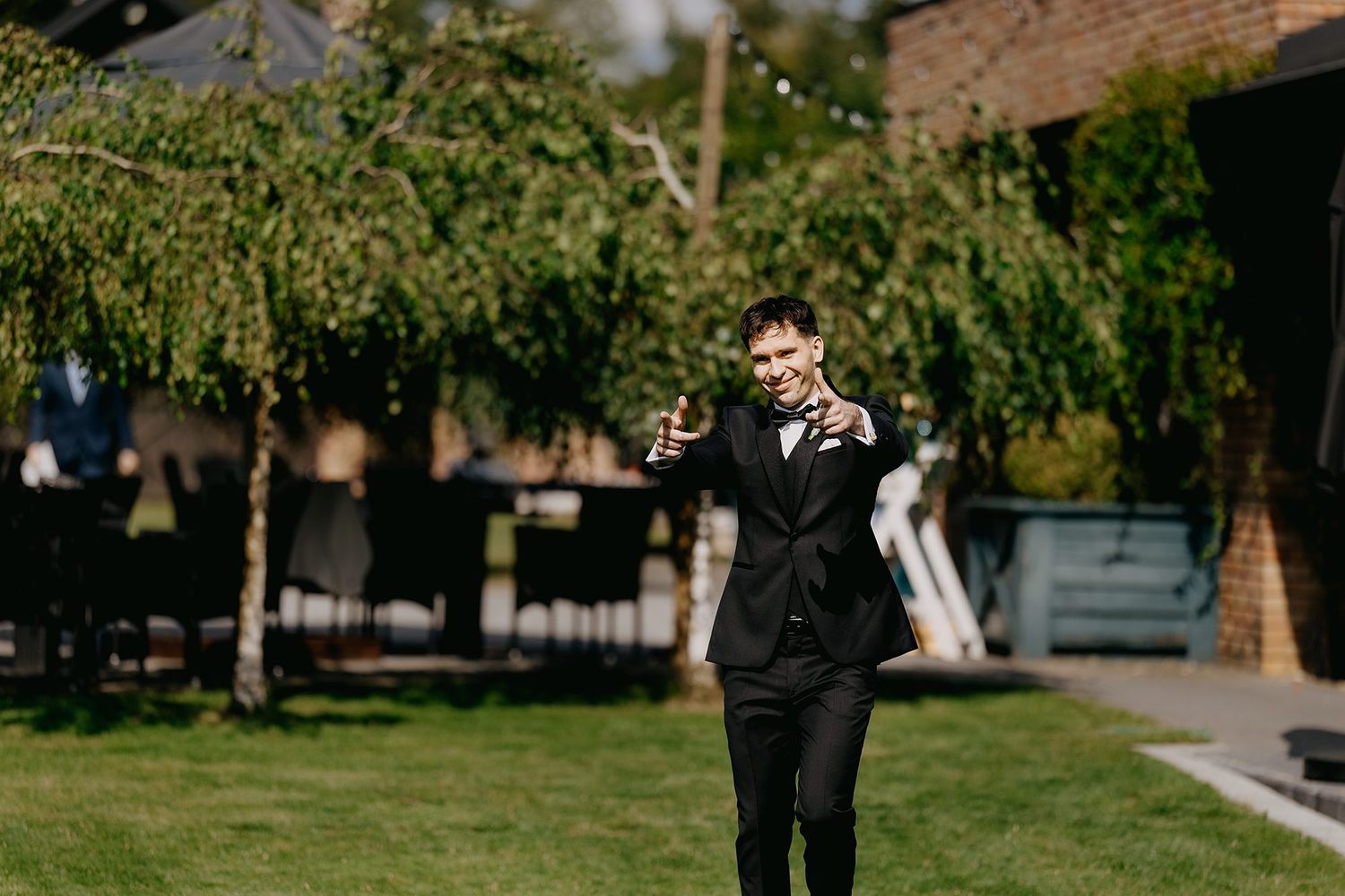 Groom in black tuxedo adjusting boutonniere on green lawn at outdoor wedding venue.