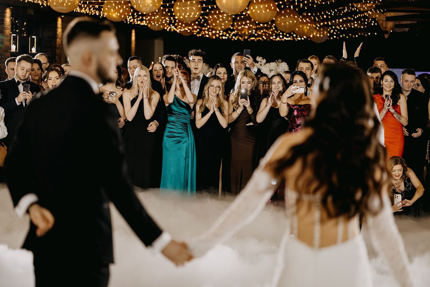 Wedding guests applaud as bride and groom dance on a fog-covered dance floor at their reception.