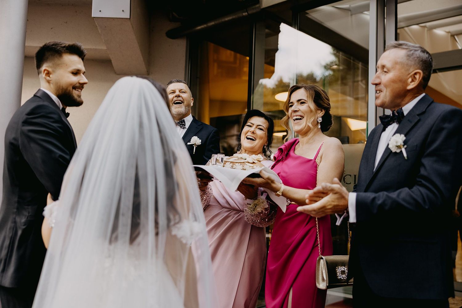 Bride and groom being greeted with bread and salt by smiling family members in pink and black formal attire.