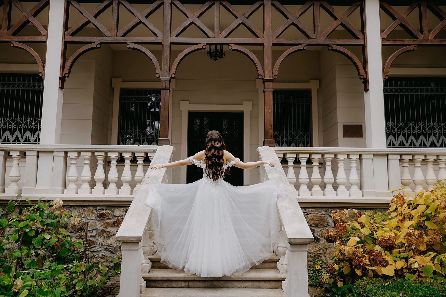 Bride in flowing white gown stands on grand staircase of elegant villa with ornate balcony.