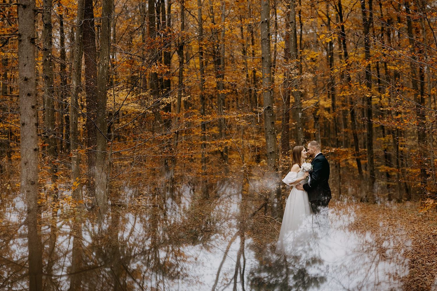 Bride and groom share romantic moment in golden autumn forest with stunning reflection below.