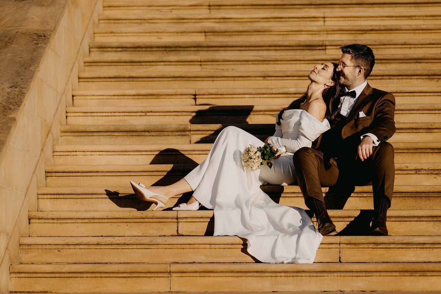 Bride and groom sit laughing together on sun-drenched stone steps in wedding attire.