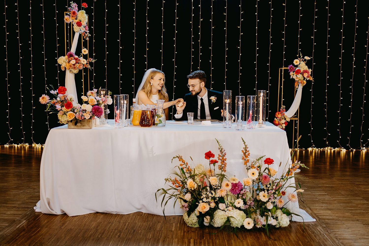 Bride and groom seated at sweetheart table adorned with colorful floral arrangements at reception.