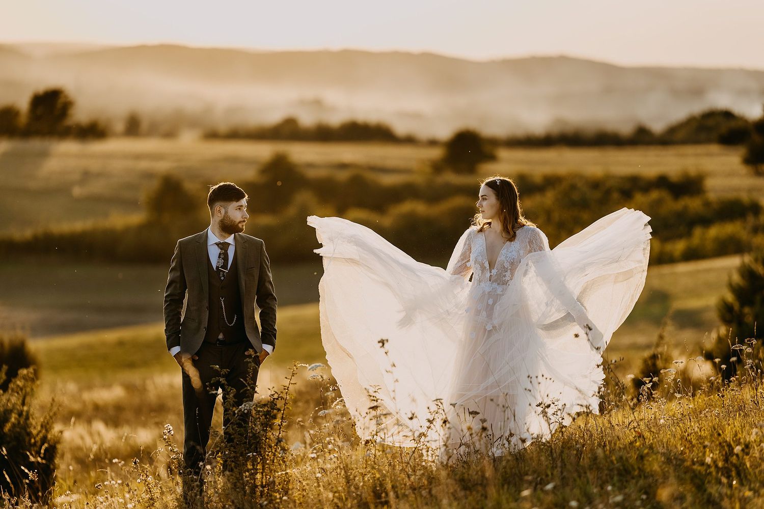 Bride and groom walking in a golden meadow with her lace dress flowing in the wind.