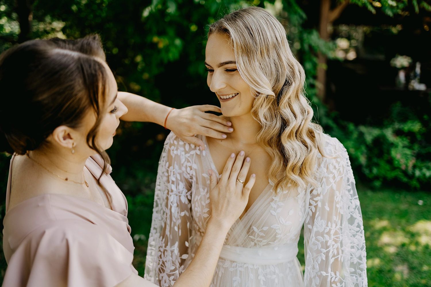 Bridesmaid tenderly adjusting bride's lace wedding dress neckline outdoors in natural light.
