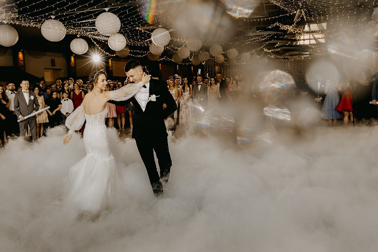 Bride and groom share first dance surrounded by dramatic fog and string lights.