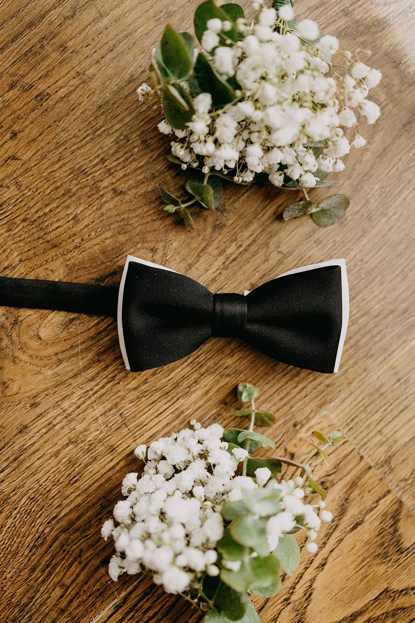 Black bow tie with white trim on wooden surface surrounded by white baby's breath flowers.