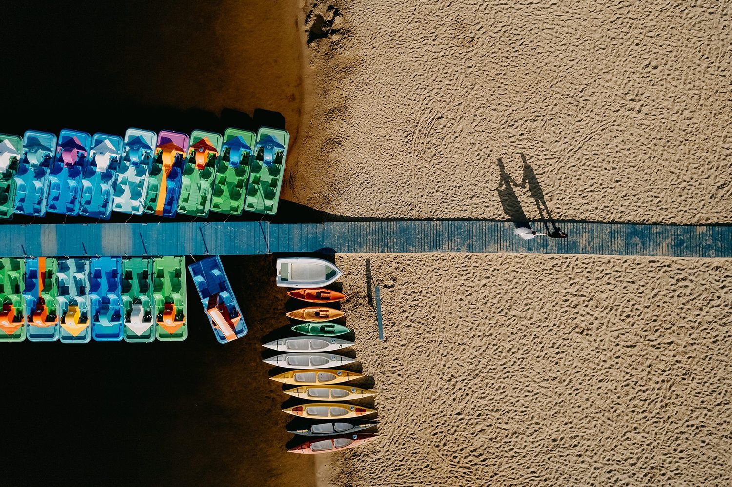 Aerial view of colorful kayaks and canoes docked at a sandy riverbank with two shadows visible.