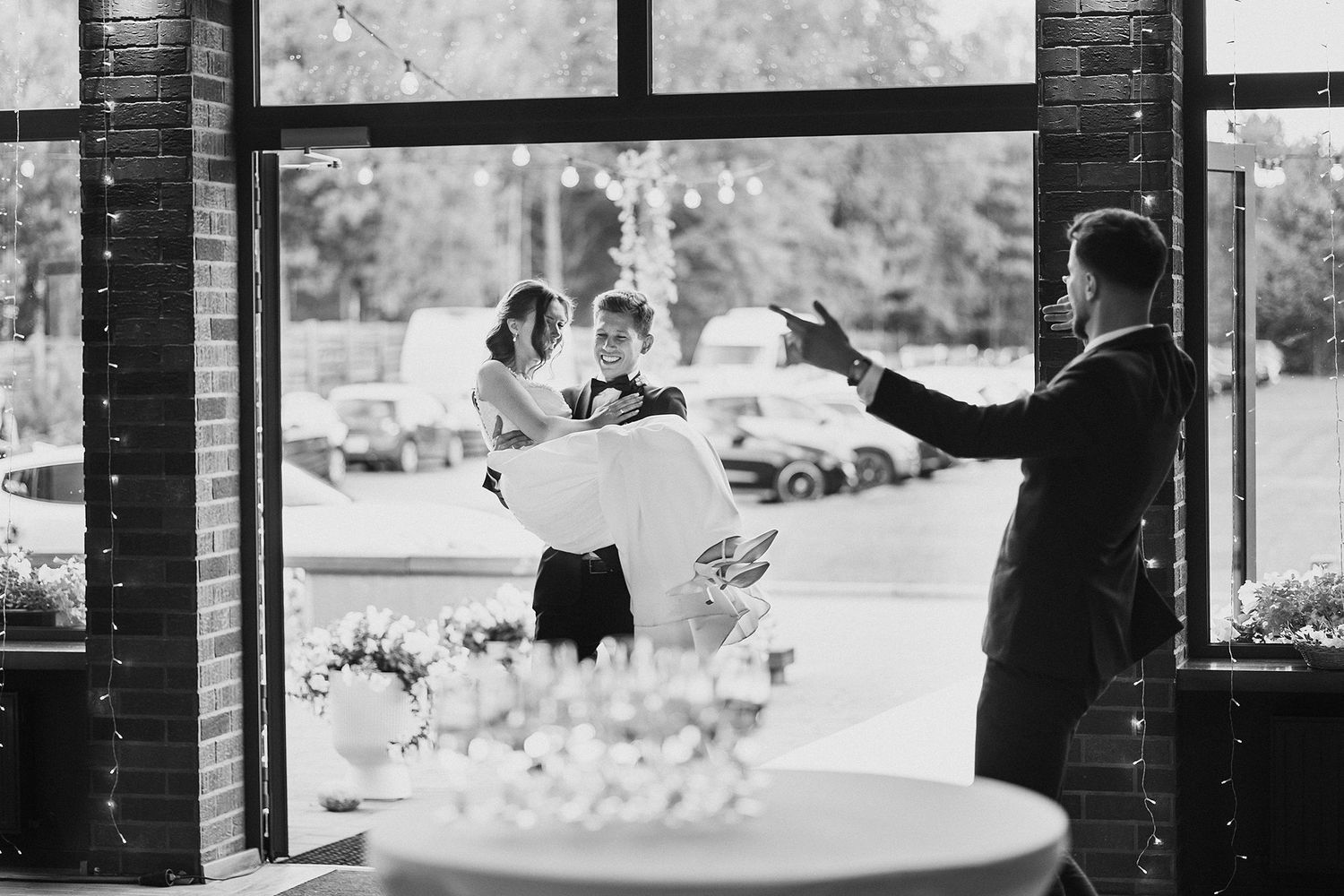 Black and white photo of groom carrying bride through a sunlit venue entrance as guest photographs them.