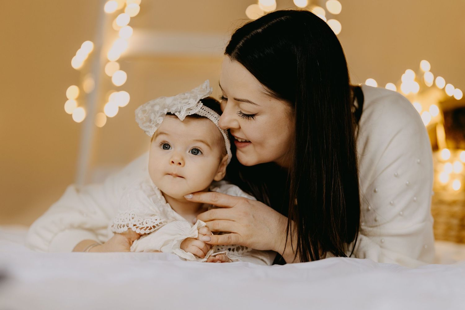 Smiling mother with long dark hair lies beside her baby girl in a white lace headband, with warm glowing string lights in the background.