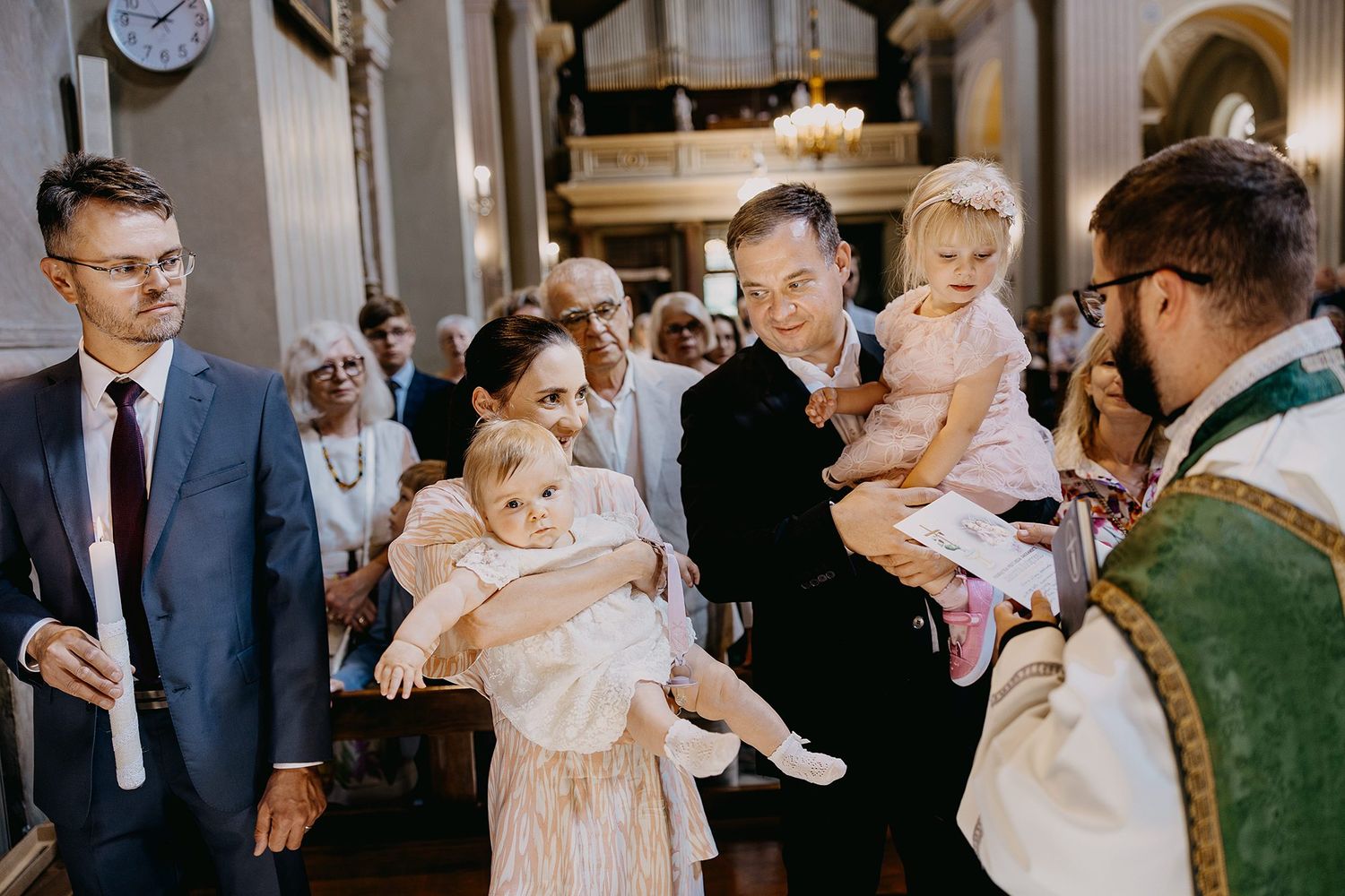 Family presenting two babies to priest during baptism ceremony in ornate church.