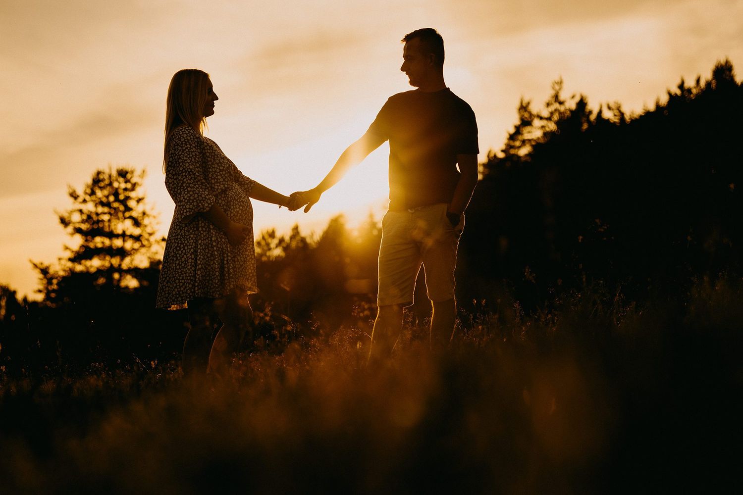 Silhouette of pregnant couple holding hands in golden sunset field with warm backlight.