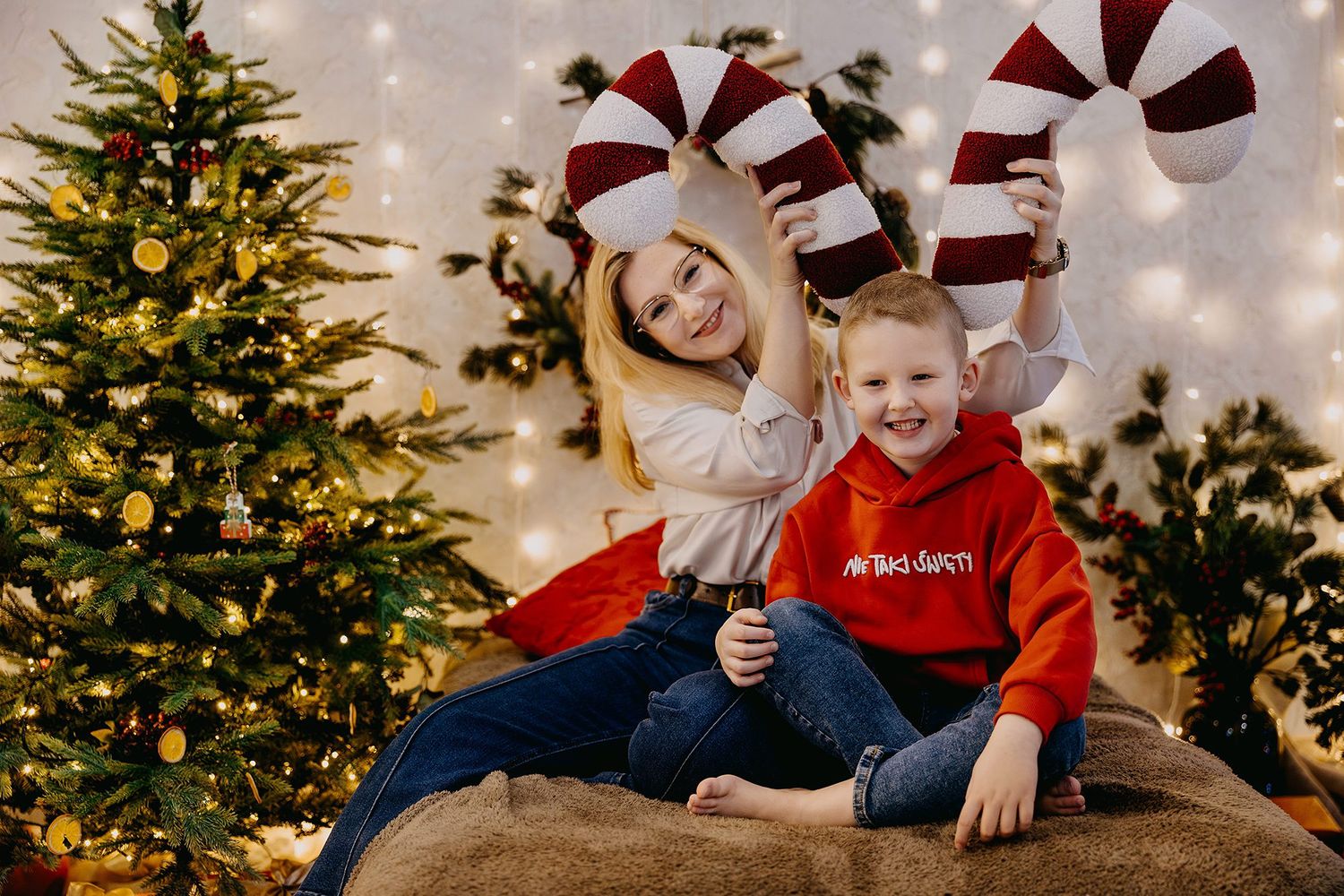 Smiling girl and young boy holding giant candy canes beside a decorated Christmas tree.